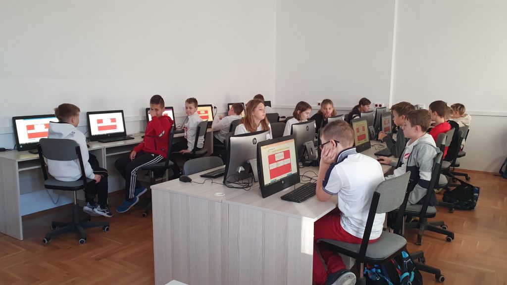 Uczniowie w sali komputerowej, siedzący przed komputerami, ubrani w barwy narodowe, rozwiązują na ekrany komputerów zadania.