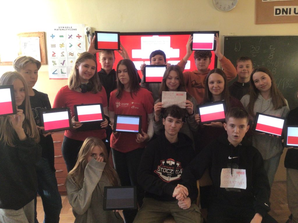 Uczniowie w strojach biało-czerwonych, trzymają w rękach tablety z wyświetloną flagą Polski, za nimi jest tablica interaktywna.