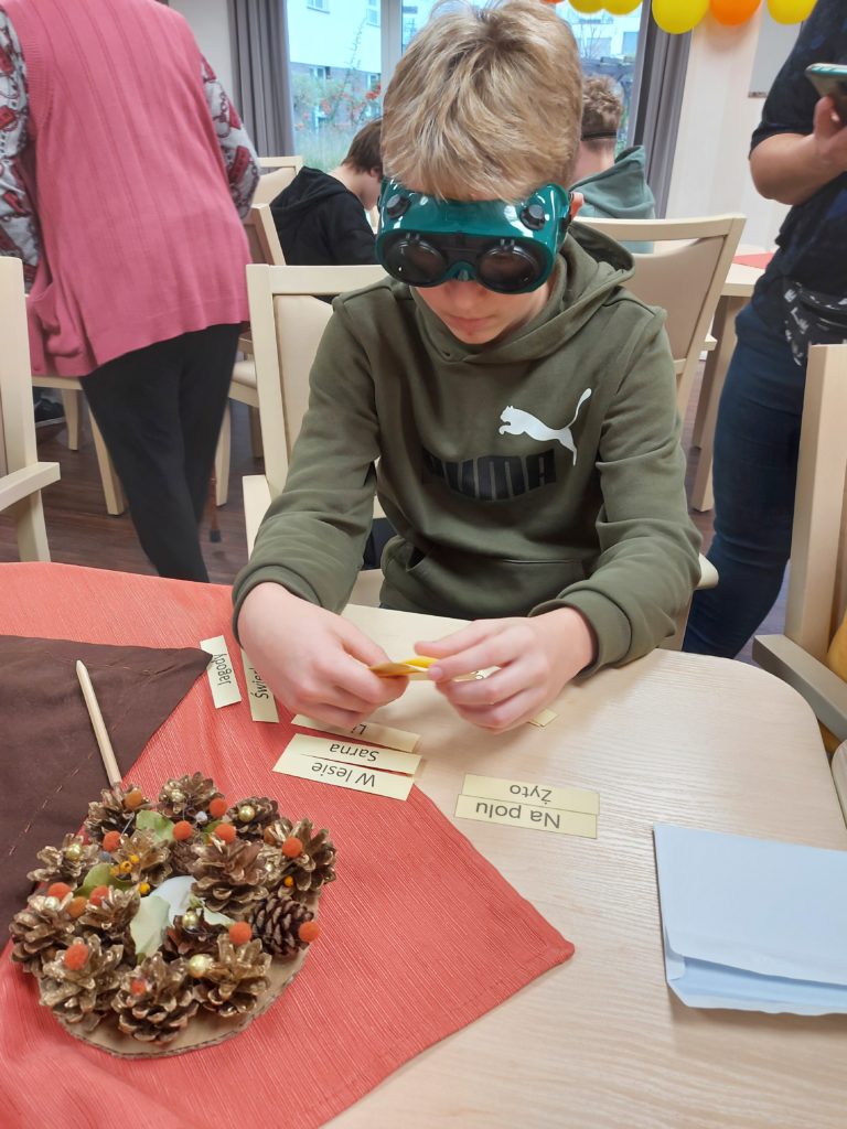Chłopiec z założonymi goglami siedzi przy stole i trzyma w ręku karteczki. Na stole znajduje się świąteczny stroik oraz leży otwarta koperta i ułożone wcześniej karteczki z napisami.