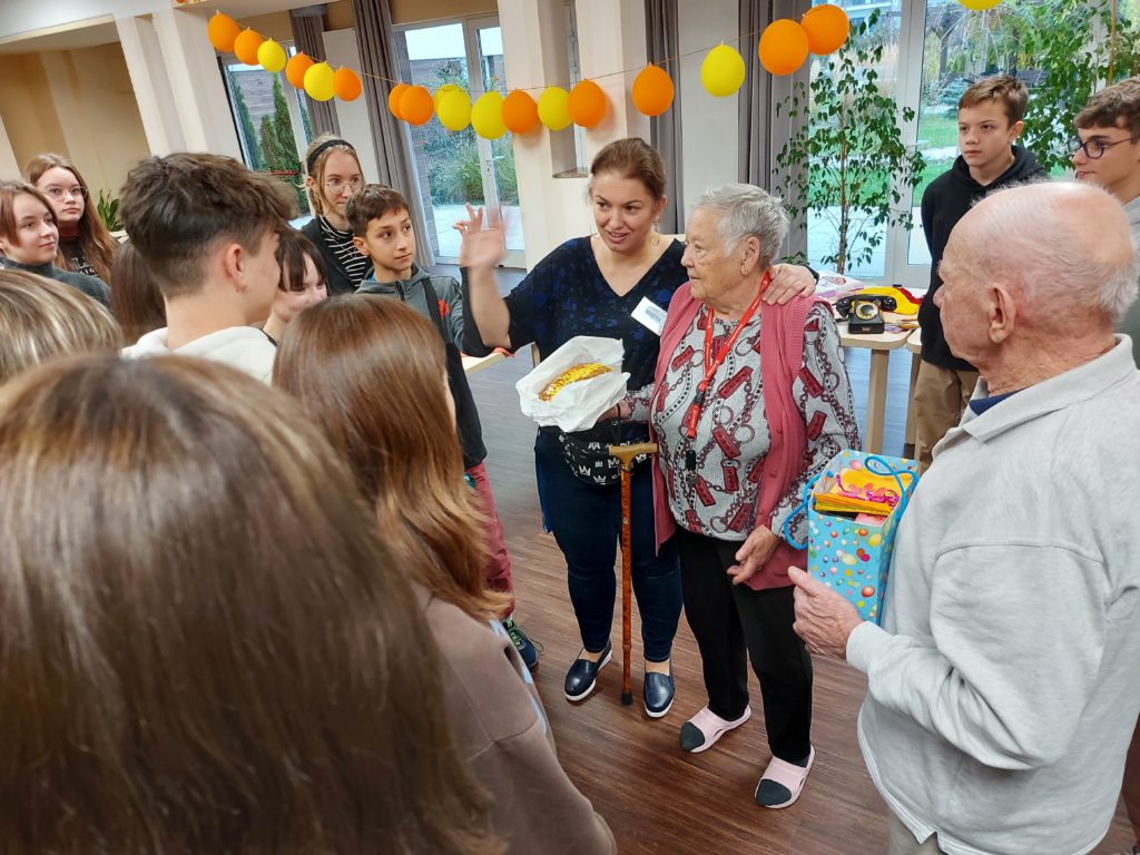 Grupa uczniów stoi wokół starszej kobiety i mężczyzny. Kobieta trzyma w ręku ciasto oraz laskę. Mężczyzna trzyma torebkę prezentową z upominkami. W tle wiszą balony.
