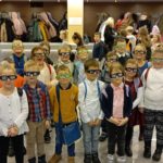 20 uczniów klasy 1c w okularach 3D stoi w dwóch szeregach, w tle widać szatnię z kurtkami oraz uczniów innej klasy.