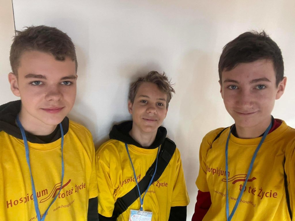 Trzech chłopców na zdjęciu typu selfie. Ubrani są w koszulki wolontariuszy, na szyjach mają powieszone identyfikatory. Na ich twarzach jest uśmiech.