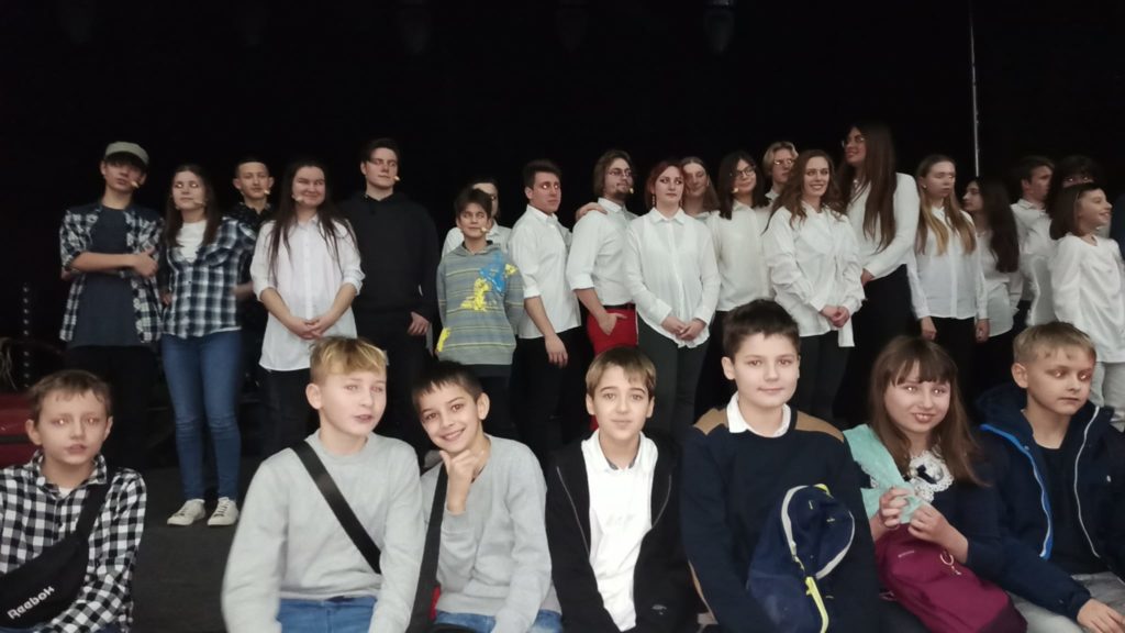 Uczniowie klas 5 siedzą w szeregu na brzegu sceny. Za nimi w szeregu stoją młodzi aktorzy, w większości ubrani w białe koszule.