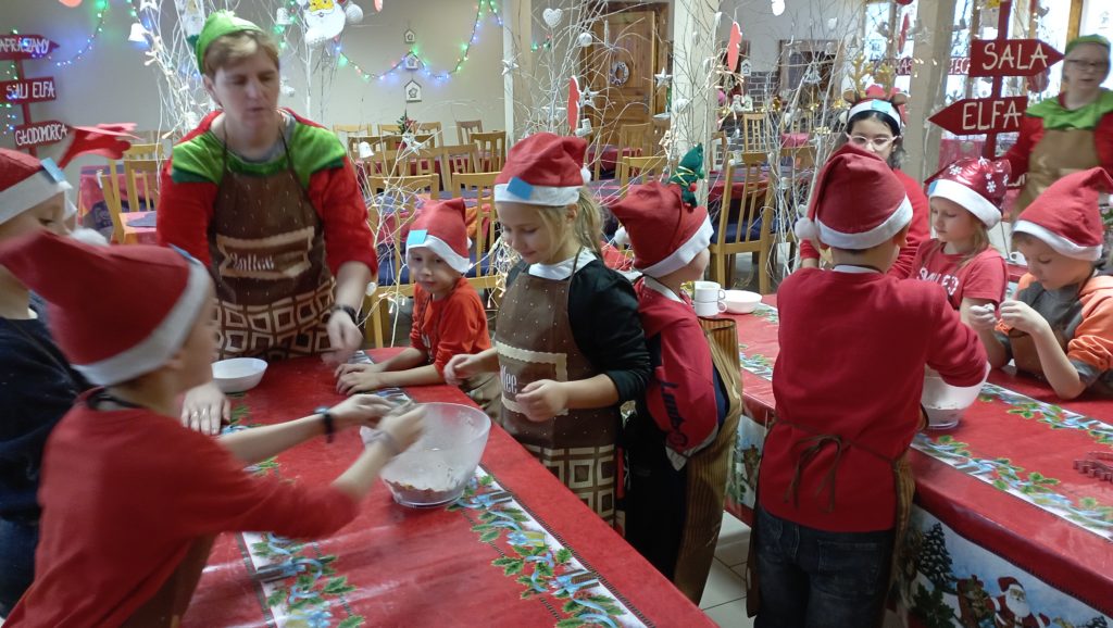 W jadalni przy dwóch stołach nakrytych obrusami stoi 9 uczniów klasy 1c w mikołajkowych nakryciach głowy oraz dwie kobiety przebrane za elfy. Dwóch uczniów trzyma ręce w miskach z ciastem piernikowym. 