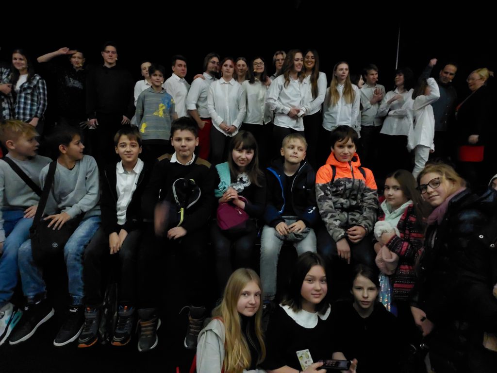 Uczniowie klas 5 siedzą w szeregu na brzegu sceny. Po prawej stronie dwie uczennice stoją przed sceną, a trzy kucają przed sceną. Z tyłu na scenie stoją aktorzy, w większości ubrani w białe koszule.