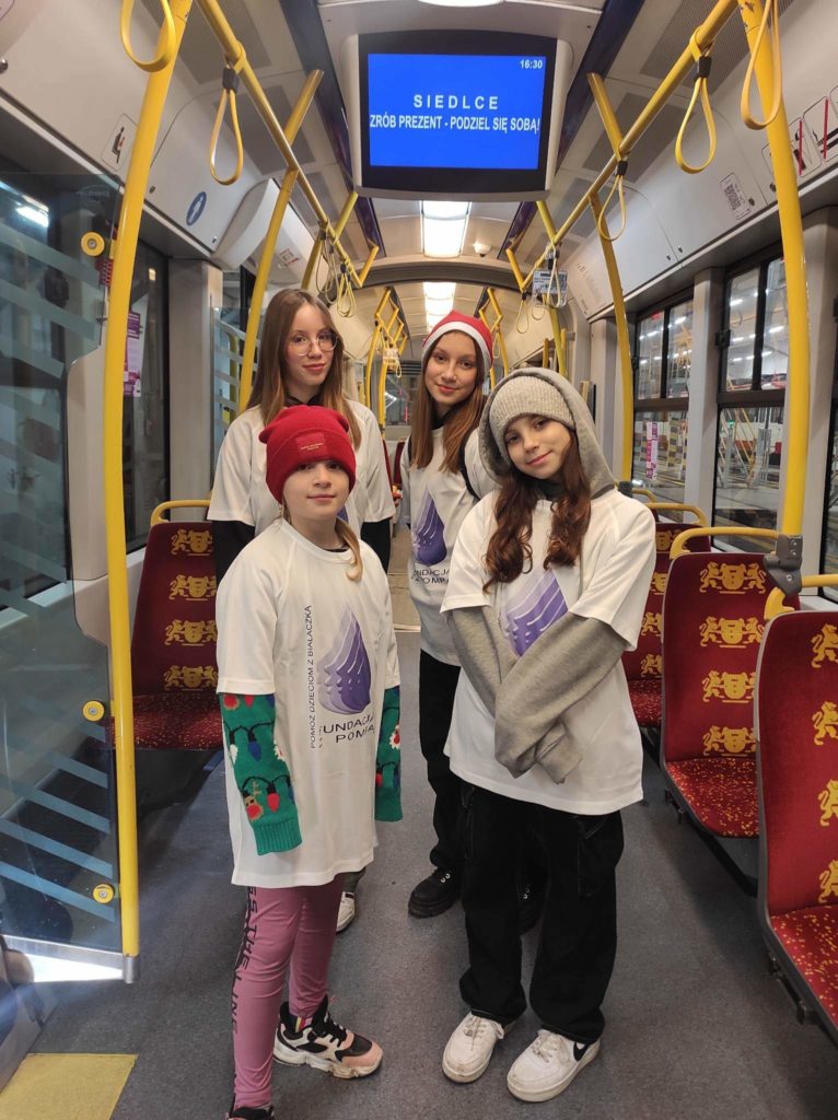 Cztery dziewczynki stoją w pustym tramwaju. Ubrane są w koszulki wolontariuszy, na szyjach mają powieszone identyfikatory. Na ich twarzach jest uśmiech.