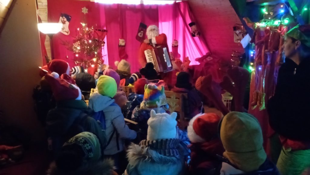 W udekorowanej sali stoi obrócona tyłem grupa uczniów klasy 1c. W tle elf i Mikołaj z akordeonem. 