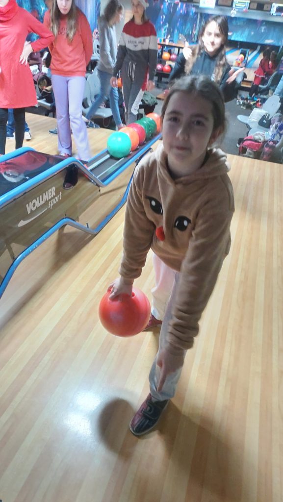 Uśmiechnięta dziewczynka ubrana w brązową bluzę z głową renifera stoi na torze bowlingowym i trzyma w rękach czerwoną kulę do gry w kręgle. W tle jest podajnik z kolorowymi kulami i stoją inne dzieci