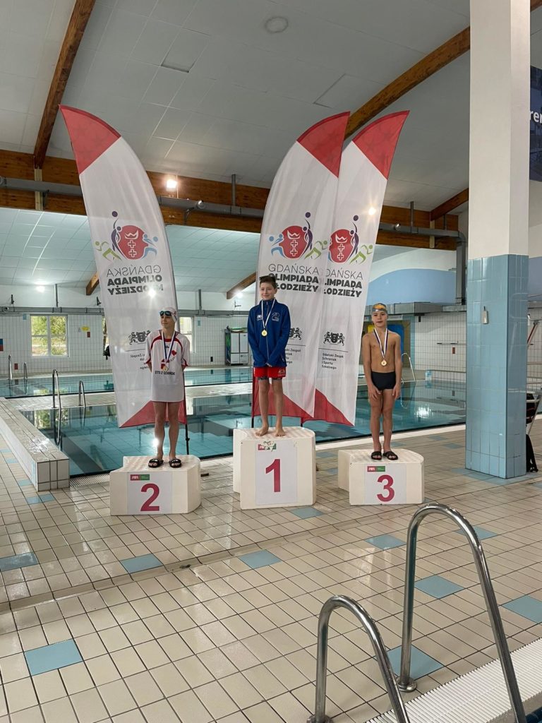 Trzech uczniów stoi na podium z medlamai na szyi a za nimi trzy flagi organizatora zawodów pływackich
