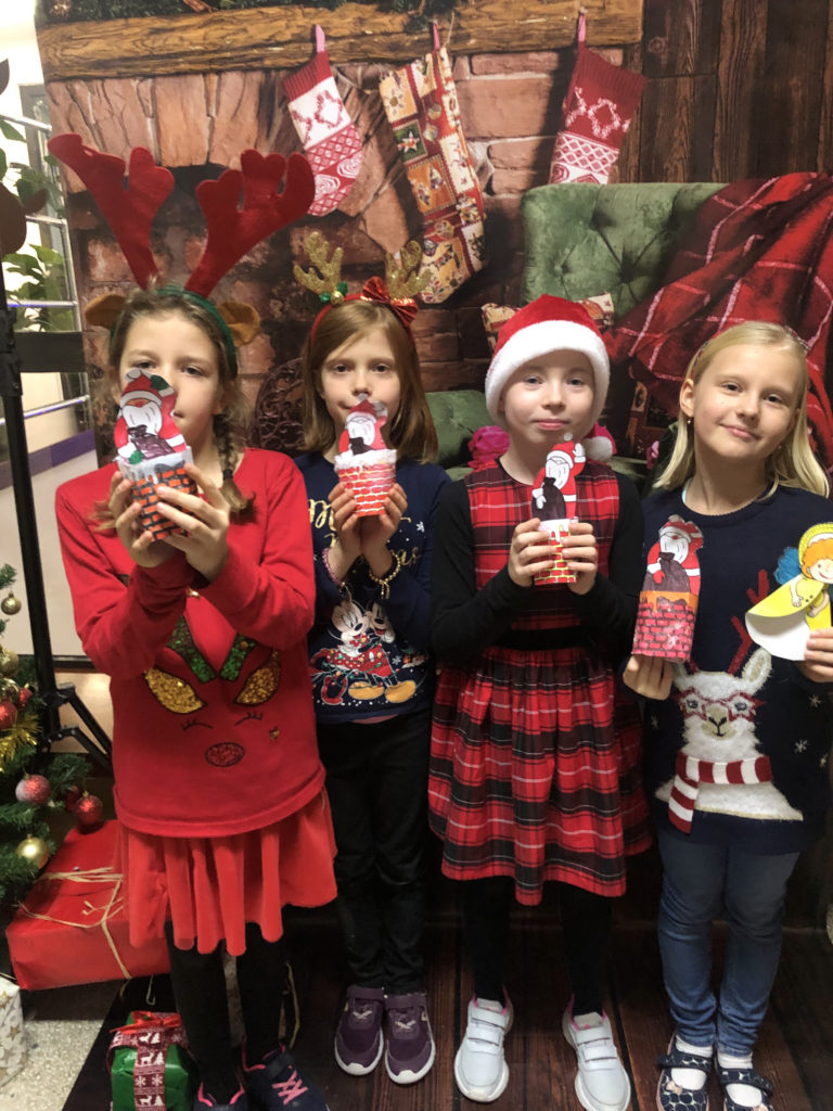 Cztery dziewczynki ubrane w świąteczne stroje trzymając figurki Mikołajów z papieru.