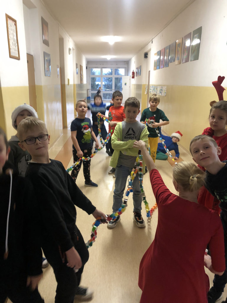 10 dzieci i jedna pani trzymają bardzo długi kolorowy papierowy łańcuch stojąc na korytarzu szkolnym