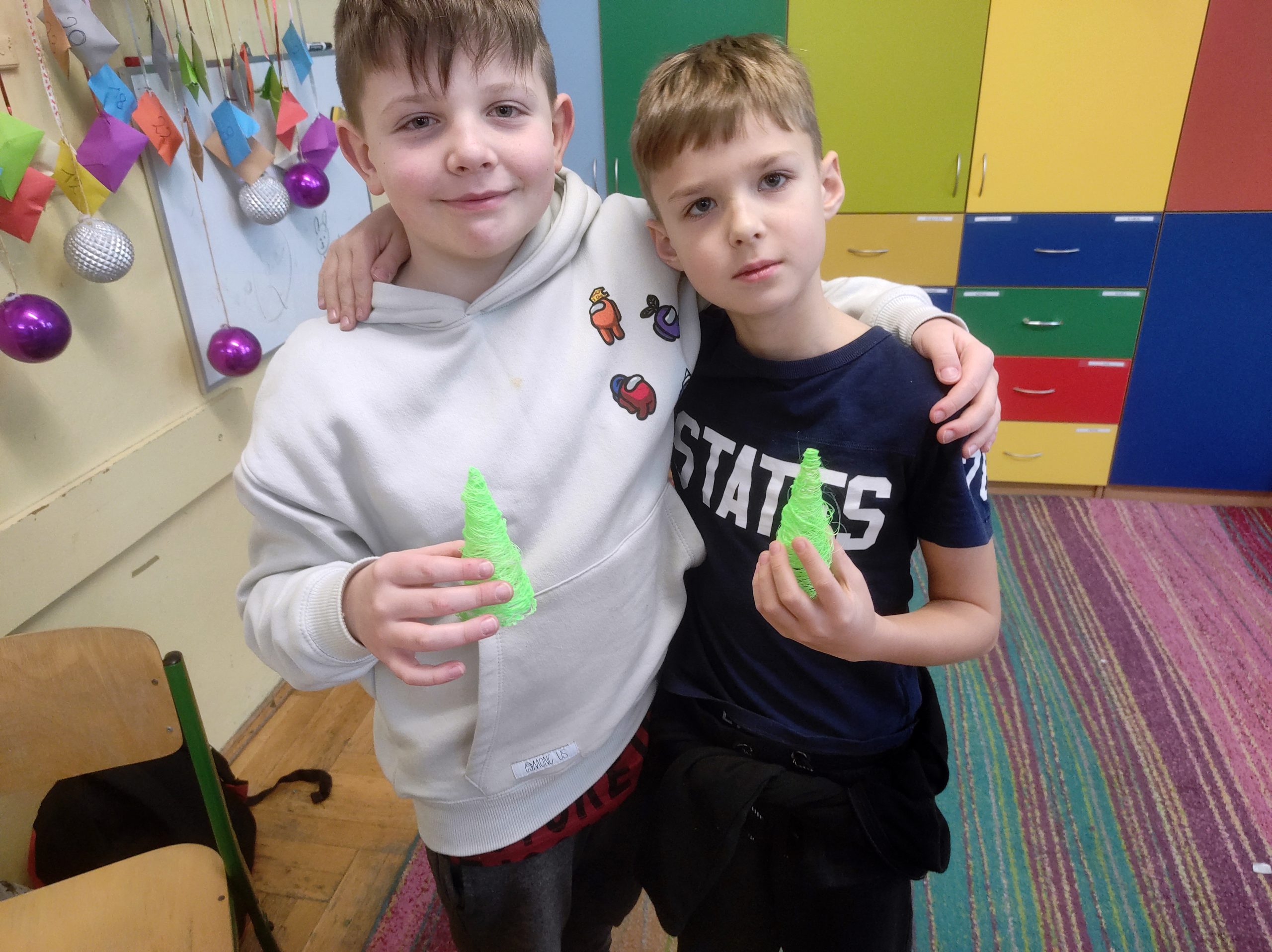 Dwóch chłopców stoi obok obejmując się. Każdy w ręku trzyma zielony stożek imitujący choinkę.
