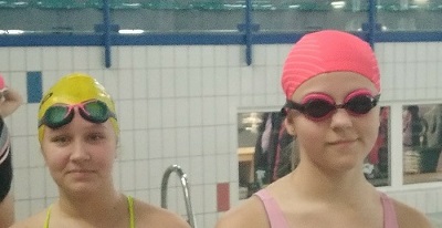Twarze dwóch zawodniczek przed startem w sztafecie pływackiej w czepkach i okularkach. Z prawej pływaczka ma je na oczach a z lewej na czole