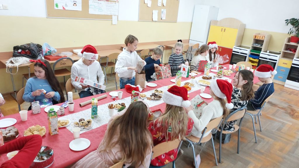 Uczniowie siedzą przy stole nakrytym czerwonym obrusem i trzymają w rękach prezenty. Na stole jest biały bieżnik, talerzyki z ciastem i ciasteczkami, kubeczki, soki w kartonikach. W tle jest sala lekcyjna z kolorowymi szafkami.