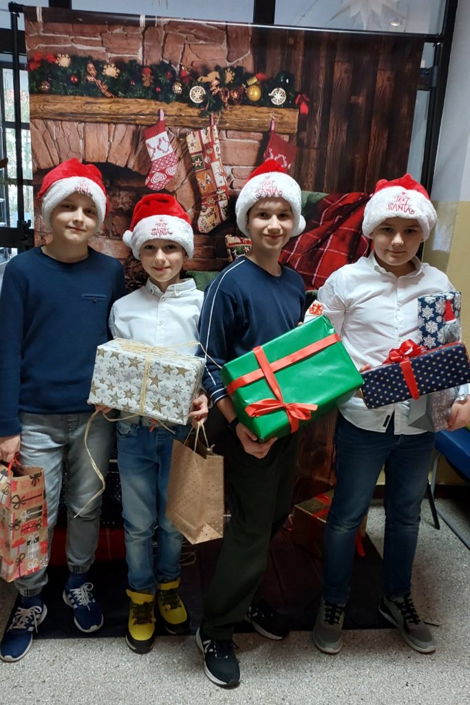Czterech uśmiechniętych chłopców ubranych w czapki św. Mikołaja stoi w szeregu i trzyma w rękach prezenty. W tle jest plakat z kominkiem i ozdobami świątecznymi.