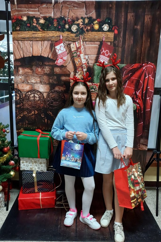 Dwie dziewczynki ubrane w spódniczki z opaskami w kształcie rogów renifera na głowach stoją obok siebie i trzymają w rękach prezenty. W tle jest plakat z kominkiem ozdobionym skarpetami świątecznymi i świerkową girlandą oraz prezenty.