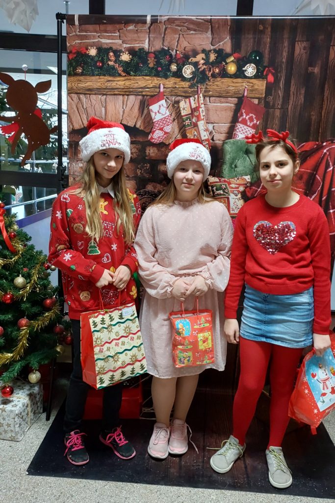 Trzy uśmiechnięte dziewczynki stoją w szeregu i trzymają w rękach prezenty. Dwie z nich mają na głowach czapki św. Mikołaja, a trzeci opaskę z rogami renifera. W tle jest plakat z kominkiem ozdobionym skarpetami świątecznymi i świerkową girlandą oraz prezenty.