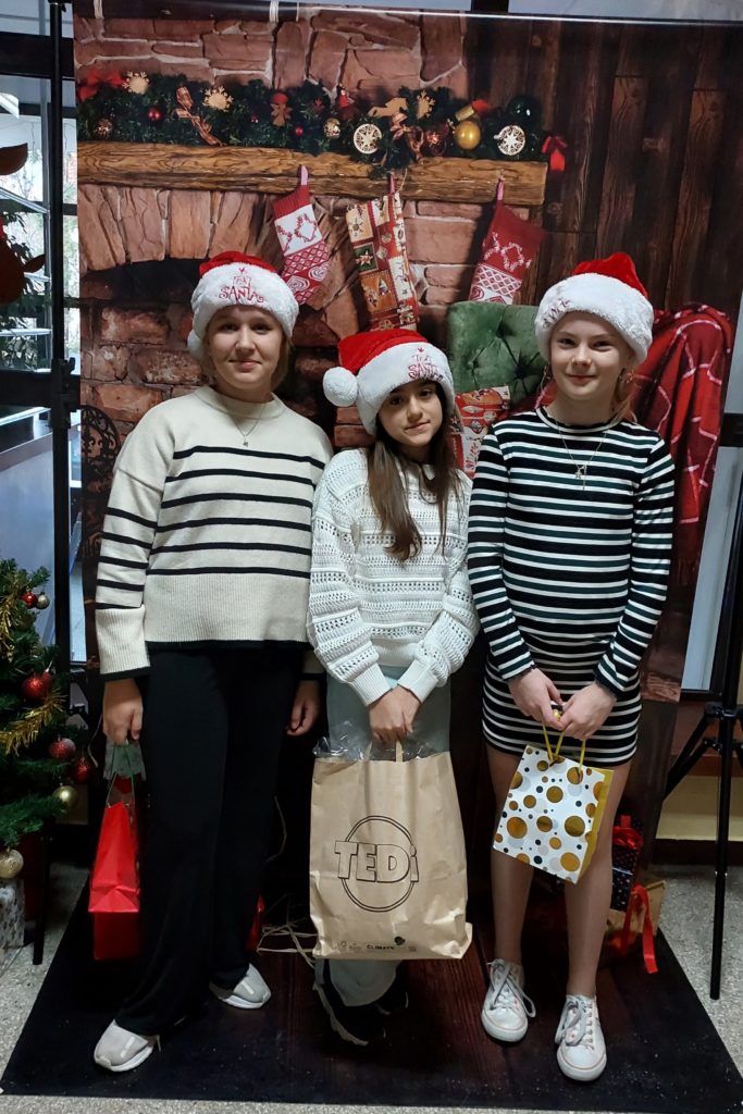 Trzy uśmiechnięte dziewczynki stoją w szeregu, ubrane w czapki św. Mikołaja. W rękach trzymają prezenty. W tle jest plakat z kominkiem ozdobionym skarpetami świątecznymi i świerkową girlandą oraz prezenty.