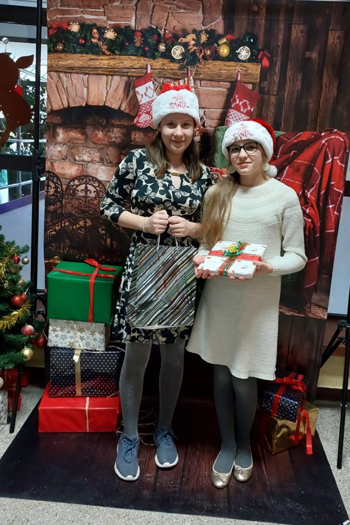 Dwie uśmiechnięte dziewczynki stoją obok siebie z czapkami św. Mikołaja na głowach. Jedna z nich ma okulary na nosie. W rękach trzymają prezenty. W tle jest plakat z kominkiem ozdobionym skarpetami świątecznymi i świerkową girlandą oraz prezenty.
