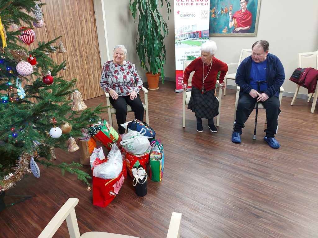 2 starsze kobiety i jeden starszy mężczyzna siedzą na krzesłach przy choince. Pod choinką leżą prezenty.