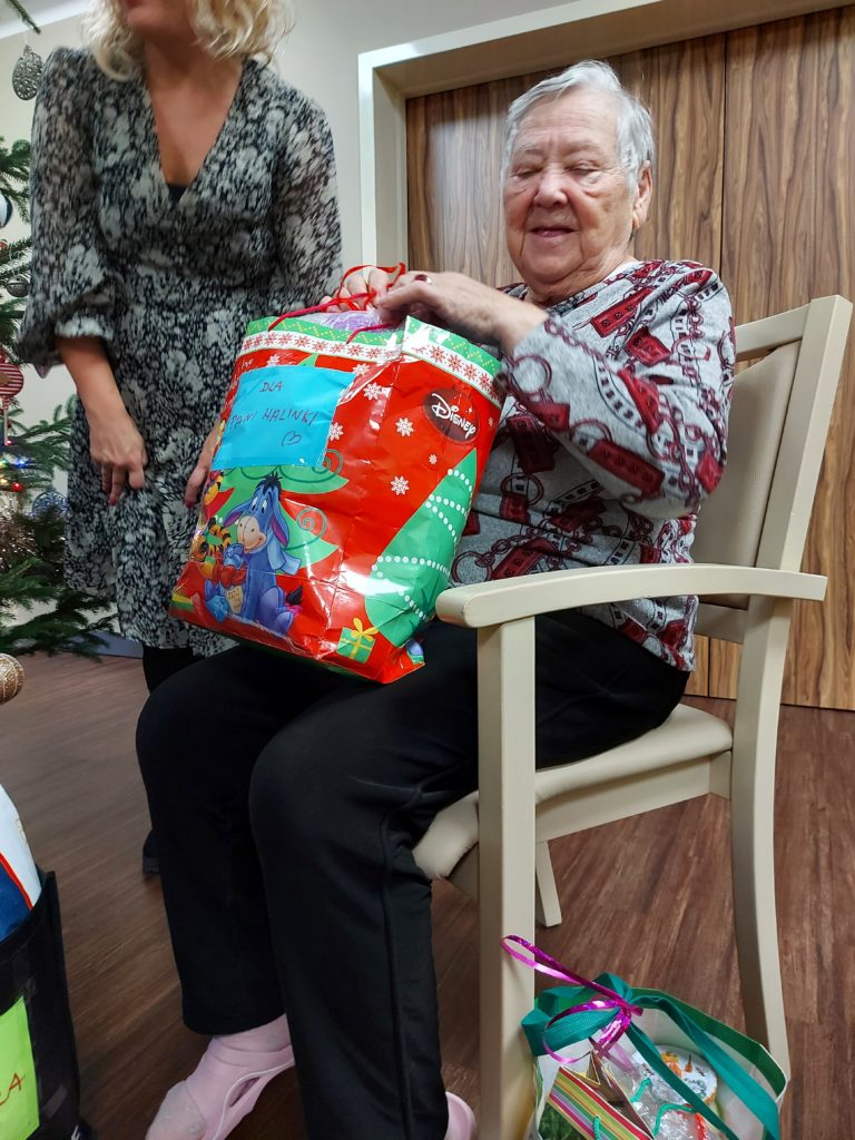 Na krześle siedzi uśmiechnięta starsza kobieta. Na kolanach trzyma dużą torbę prezentową. Torba z prezentem leży także przy krześle. Obok siędzącej staruszki stoi kobieta.