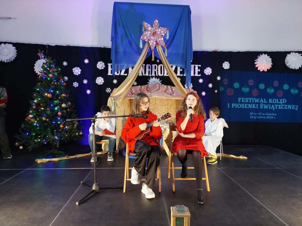 Dwie dziewczynki siedzą na krzesłach na scenie. Jedna trzyma w ręku mikrofon śpiewając do niego. Druga gra na ukulele, a przy niej stoi statyw z mikrofonem. Za nimi siedzi dwóch chłopców ubranych w strój anioła.