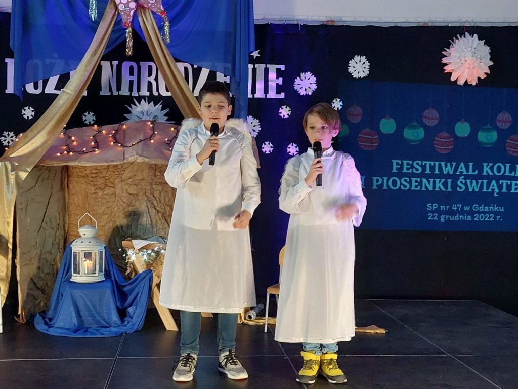 2 chłopców ubranych w strój anioła stoją na scenie i trzymają w ręku mikrofony. Za nimi stoi szopka i żłóbek.