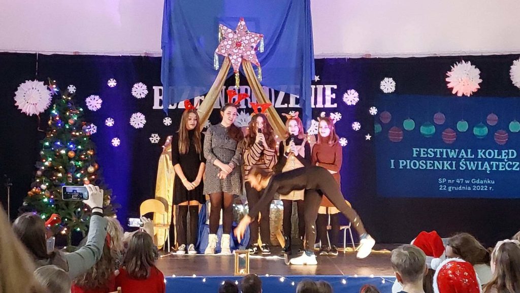 5 dziewczyn stoi na scenie - dwie z nich trzymają w ręku mikrofony. Przed nimi jedna dziewczyna wykonuje skłon chcąc zrobić gwiazdę. Przed sceną jest widownia, na której siedzą dzieci.