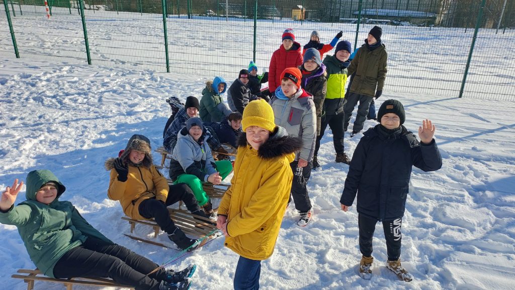 Kilkanaścioro uśmiechniętych chłopców ubranych w zimowe, kolorowe kurtki i czapki stoi na boisku szkolnym, przed nimi na sankach siedzą chłopcy. Na boisku jest dużo śniegu.