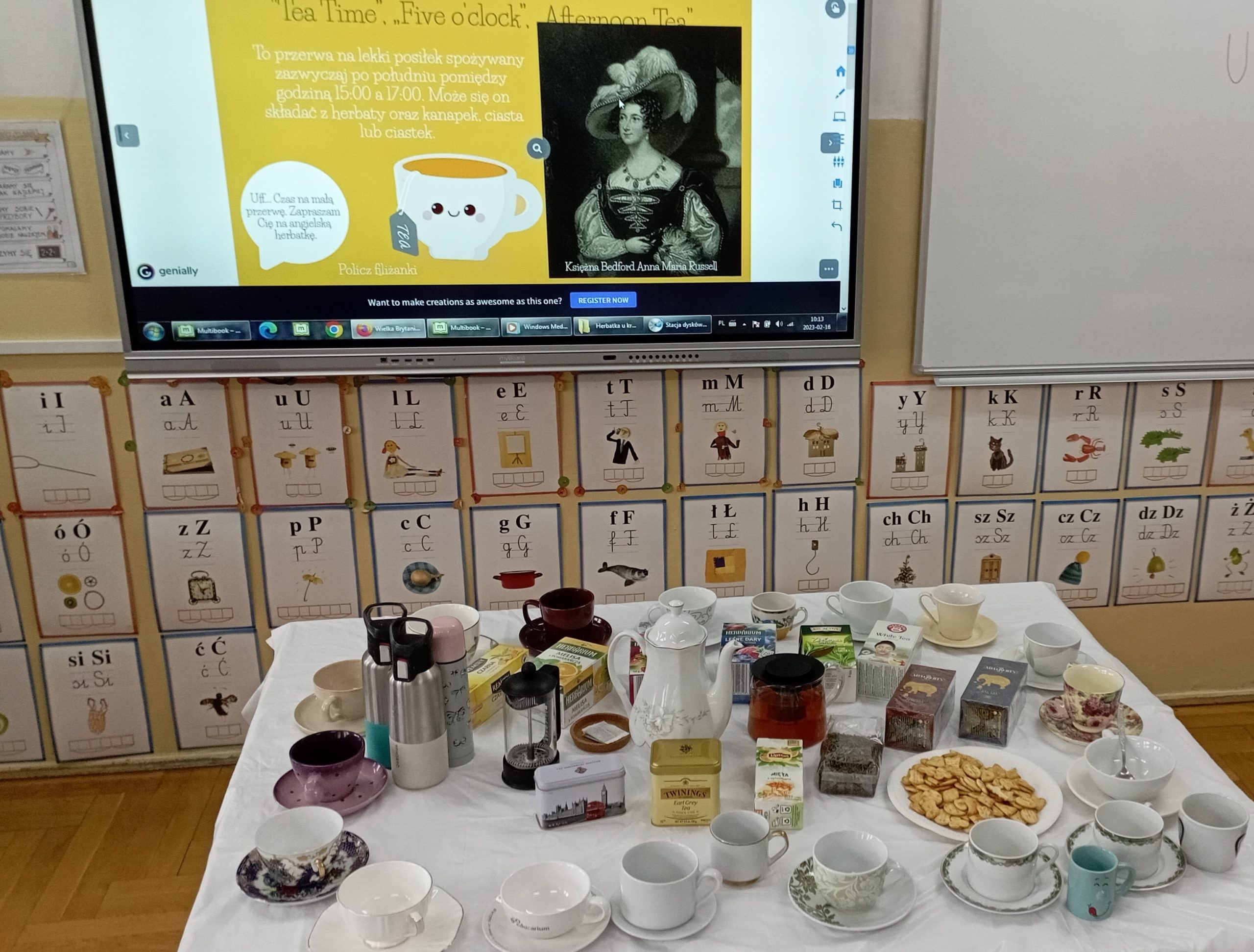 Na tle tablicy multimedialnej wyświetlającej slajd w prezentacji na temat zwyczaju picia herbaty stoi stół przykryty białym obrusem, a na nim stoją filiżanki, dzbanki i opakowania herbat.