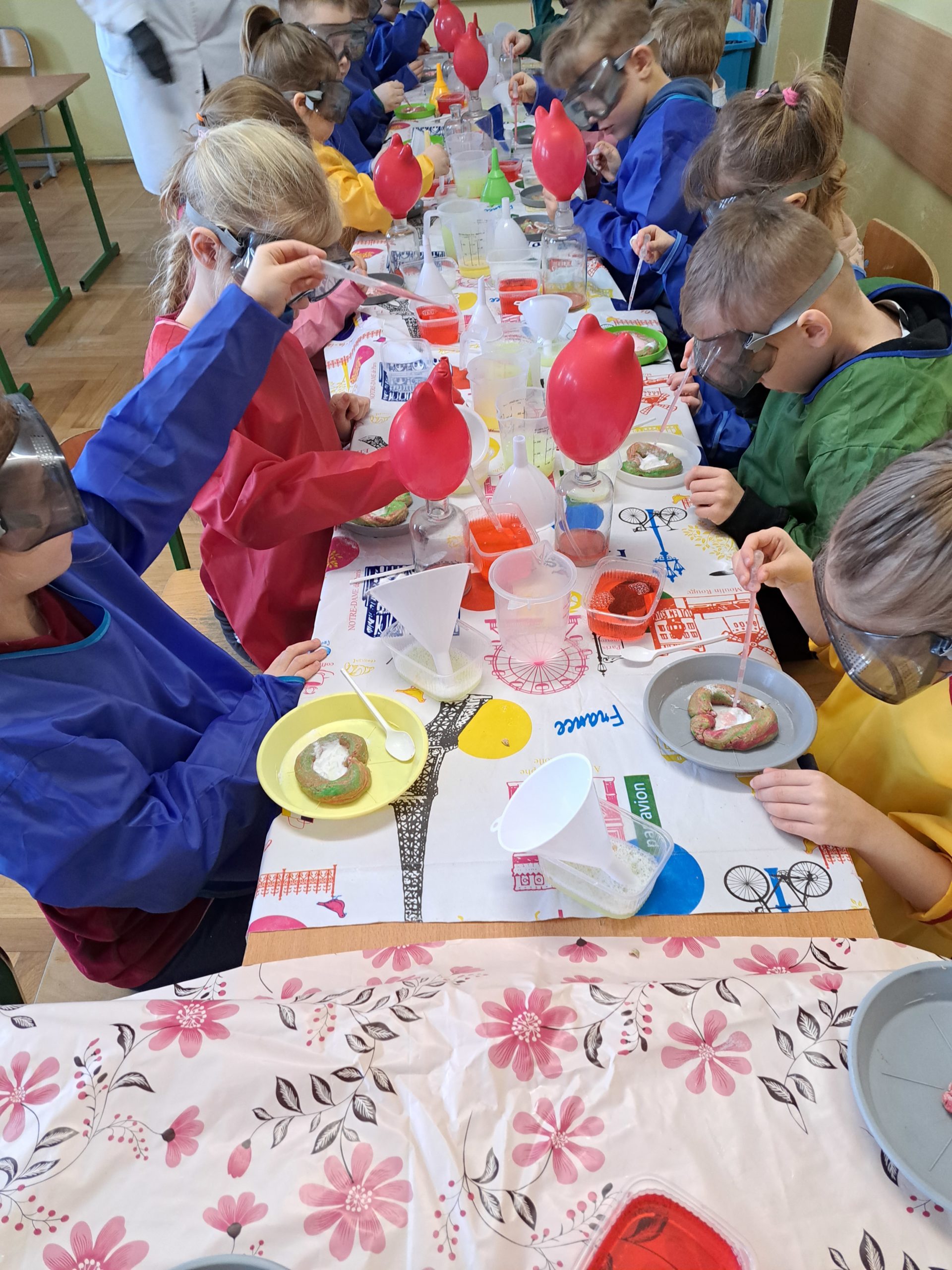 Grupa dzieci siedzi przy stole w kolorowych fartuchach i ochronnych okularach. Na kolorowych plastikowych talerzykach leżą ulepione serca, które są wypełnione specjalnym proszkiem. Dzieci polewają ten proszek cieczą za pomocą pipetki. Z proszku powstaje kolorowy wulkan, który wypływa poza serce. 