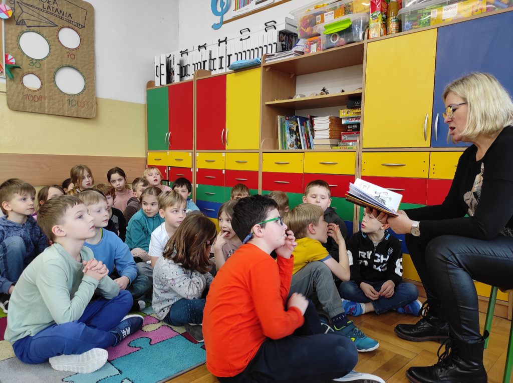 Nauczyciel czyta ksiażkę. Na dywanie w klasie siedzą dzieci, słuchając czytanego tekstu