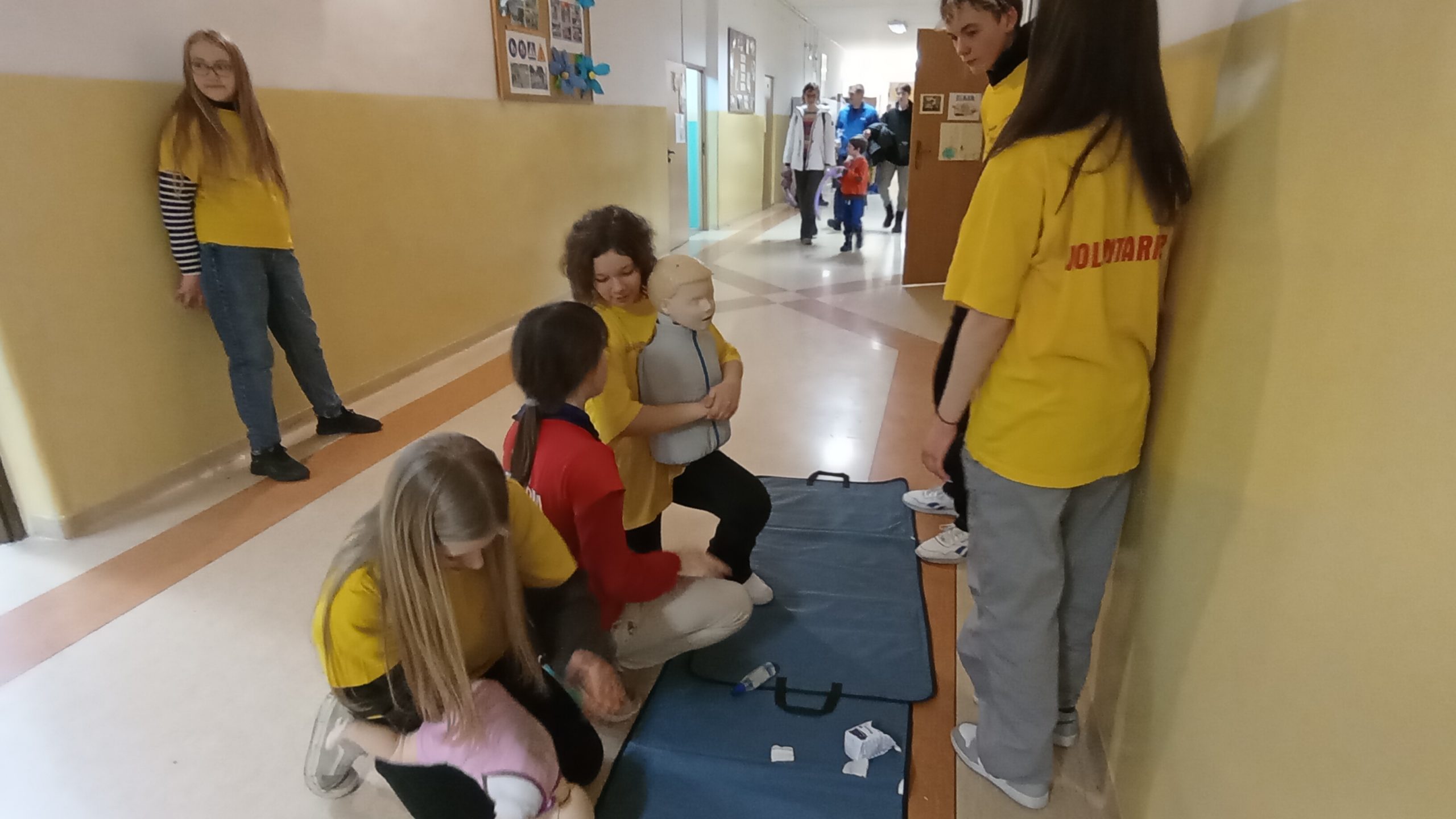 Na szkolnym korytarzu dwie uczennice w żółtych koszulkach klęczą ćwicząc odkrztuszanie dzieci na fantomach, jedna uczennica w czerwonej koszulce kuca przy nich, 3 uczniów się przyglada. W tle uczestnicy Dnia Otwartego. 