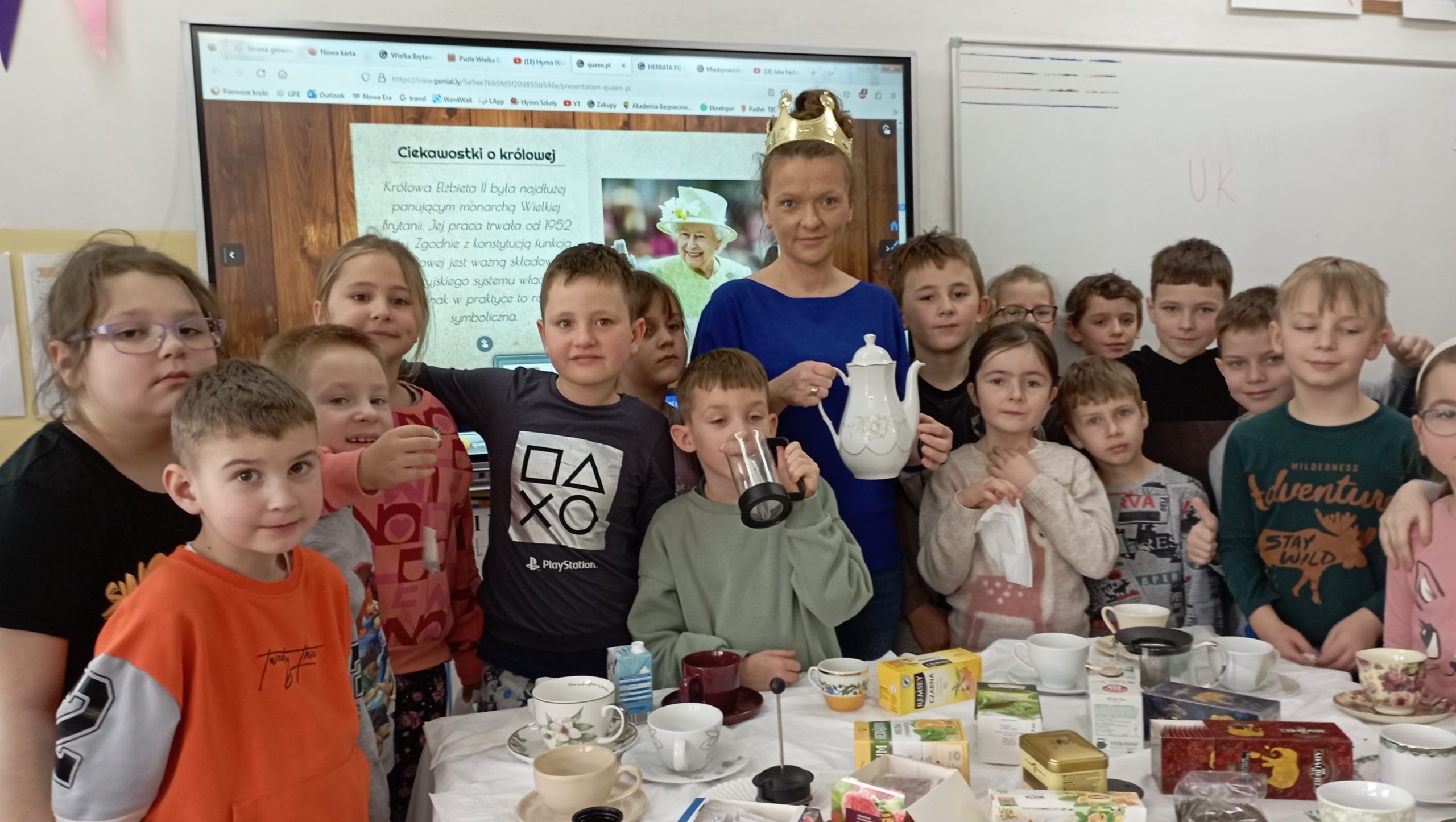 Za stołem z filiżankami i herbatami stoją uczniowie wraz nauczycielką z koroną na głowie i imbrykiem w dłoni. W tle tablica multimedialna ze zdjęciem królowej Elżbiety II