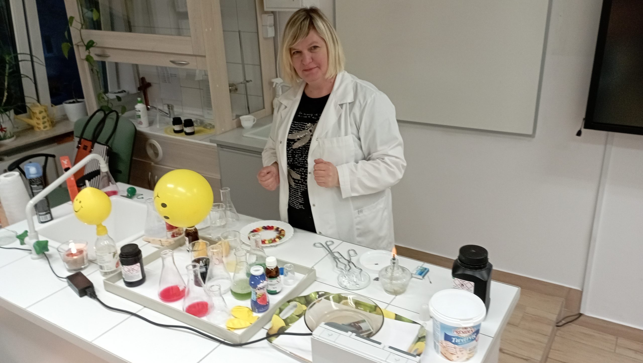 W szkolnej pracowni chemicznej na stole roboczym stoją menzurki, zlewki, preparaty i narzędzia w trakcie eksperymentów. Na pierwszym planie nauczycielka chemii prowadząca eksperymenty. W tle tablica.