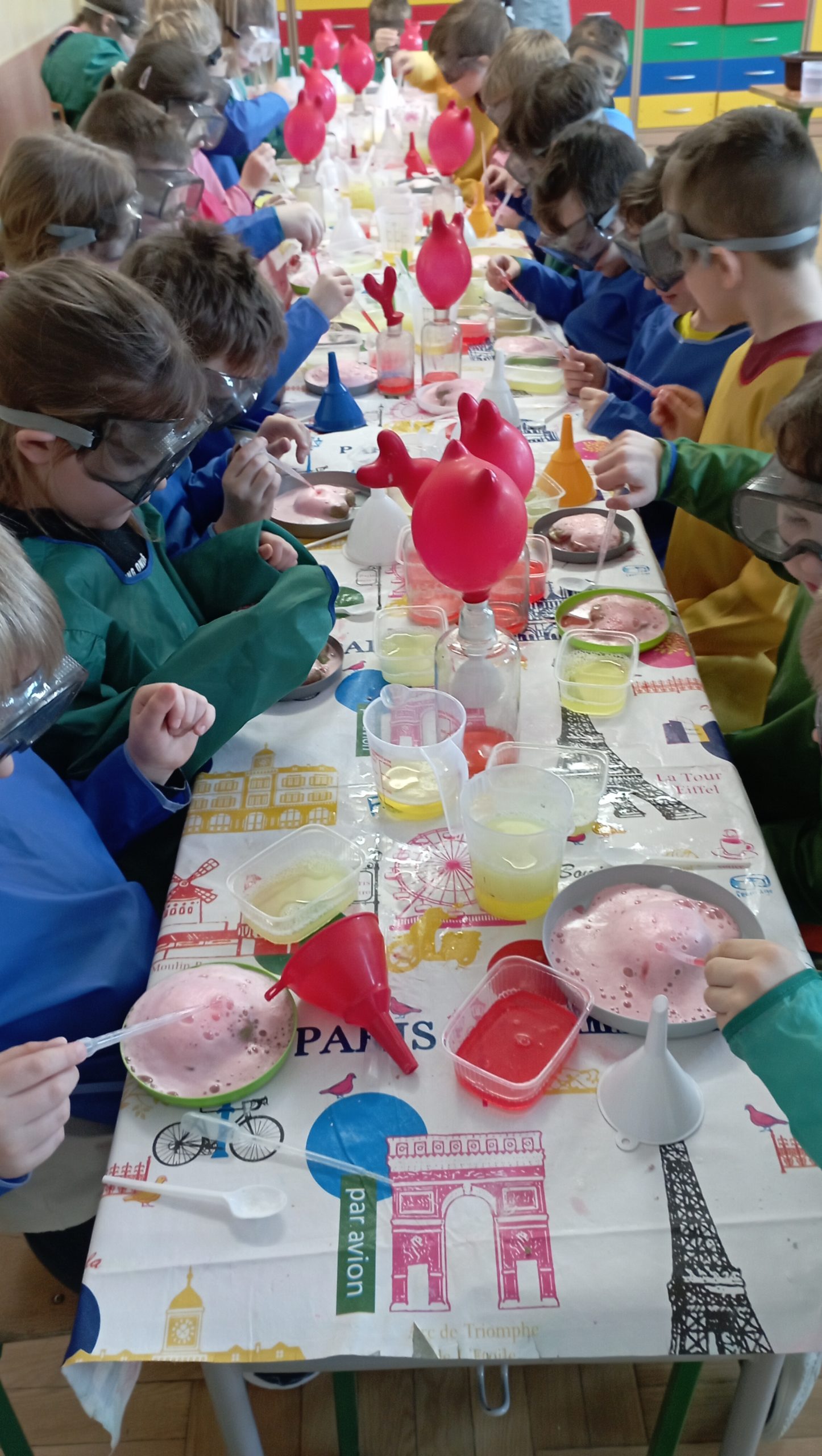 Grupa dzieci siedzi przy stole w kolorowych fartuchach i ochronnych okularach. Na plastikowych talerzykach powstają kolorowe pieniące się wulkany, dzieci polewają je płynem z pipetki.