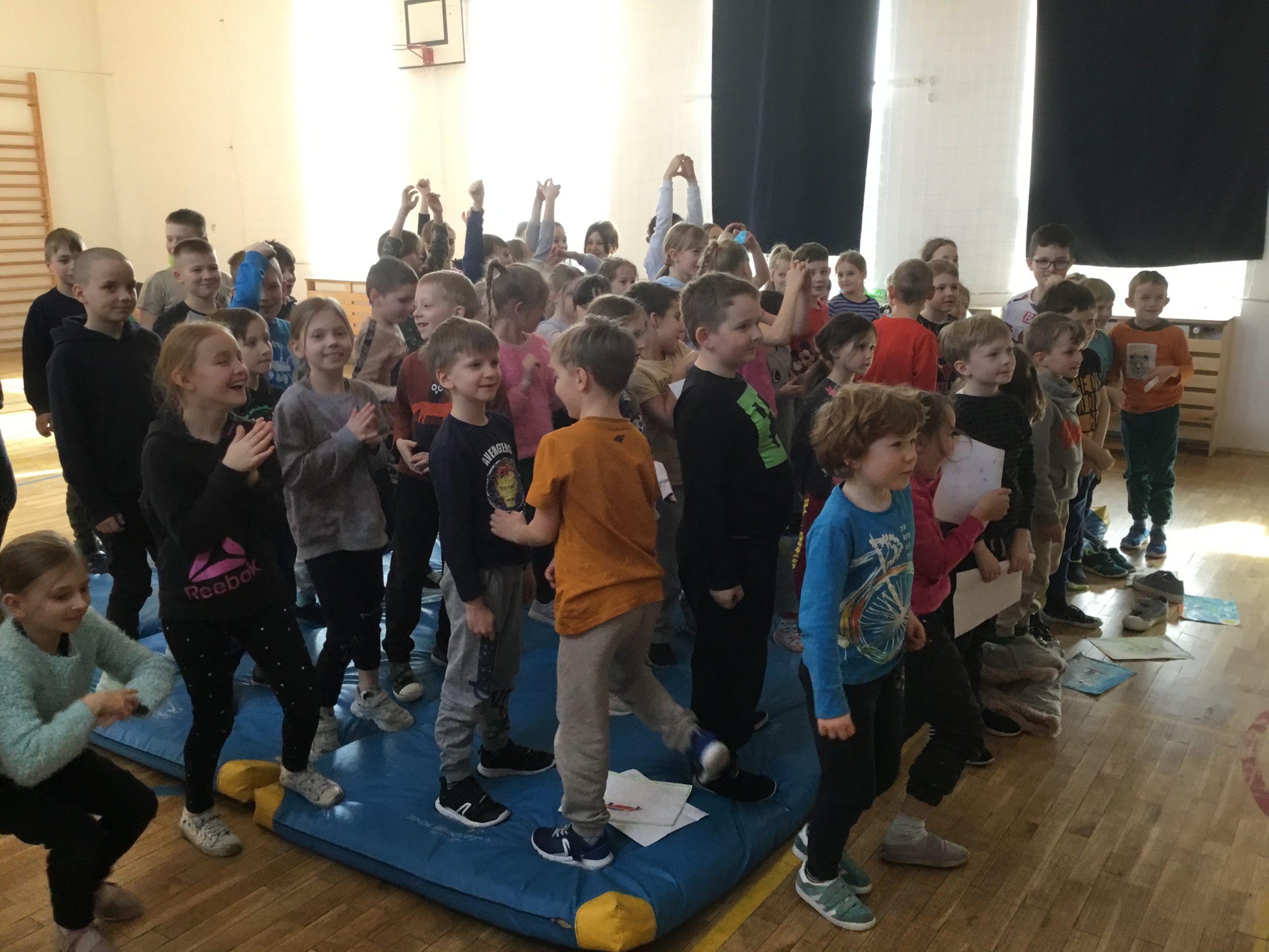 dzieci z klas młodszych na sali gimnastycznej podczas spotkania autorskiego zgłaszające się do odpowiedzi