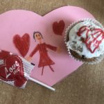 Kartka walentynkowa przestawiający dziewczynkę i serduszka. Na niej leży lizak w kształcie serca i muffinka z lukrowany czerwonym sercem.