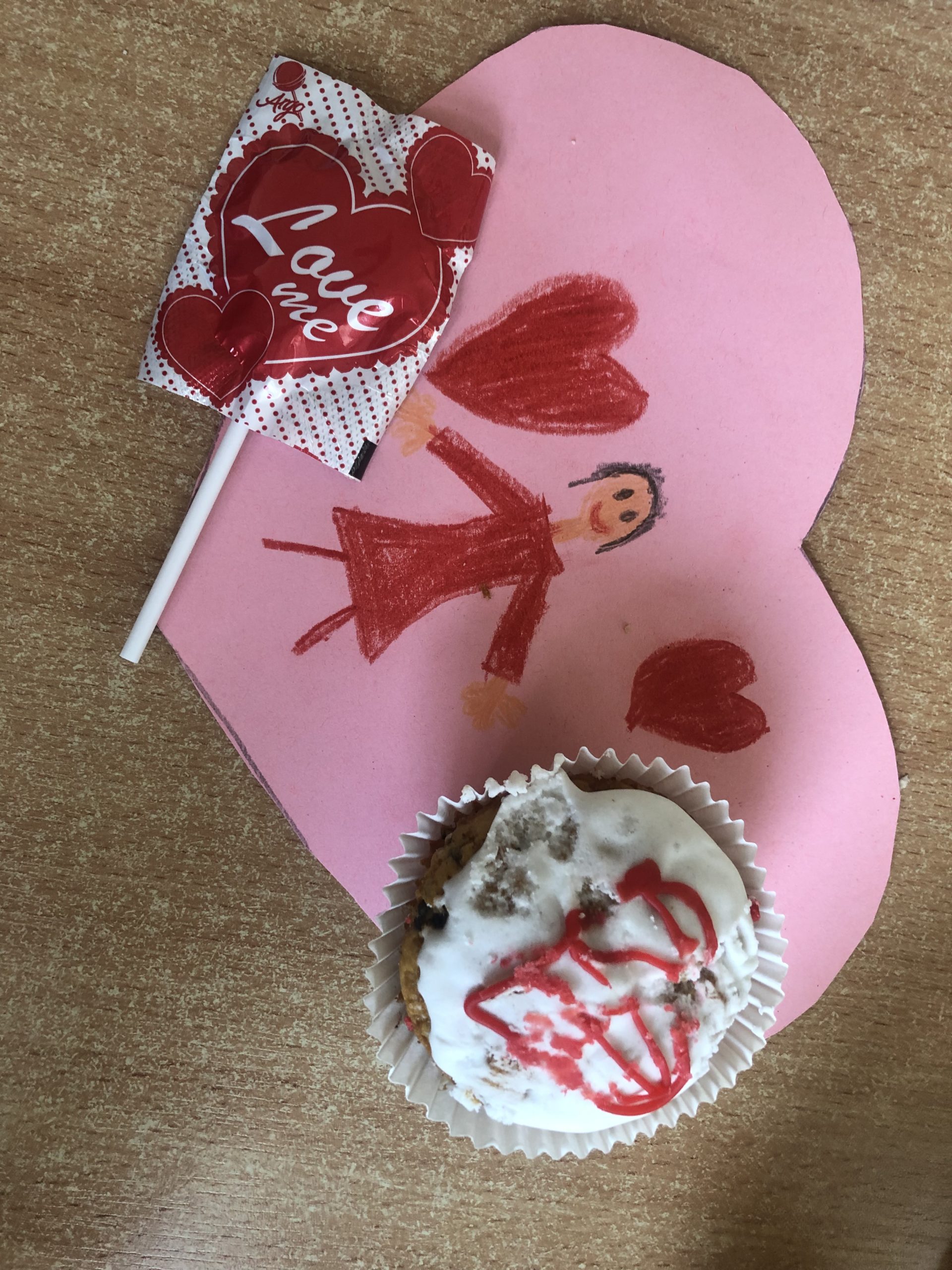 Kartka walentynkowa przestawiający dziewczynkę i serduszka. Na niej leży lizak w kształcie serca i muffinka z lukrowany czerwonym sercem.