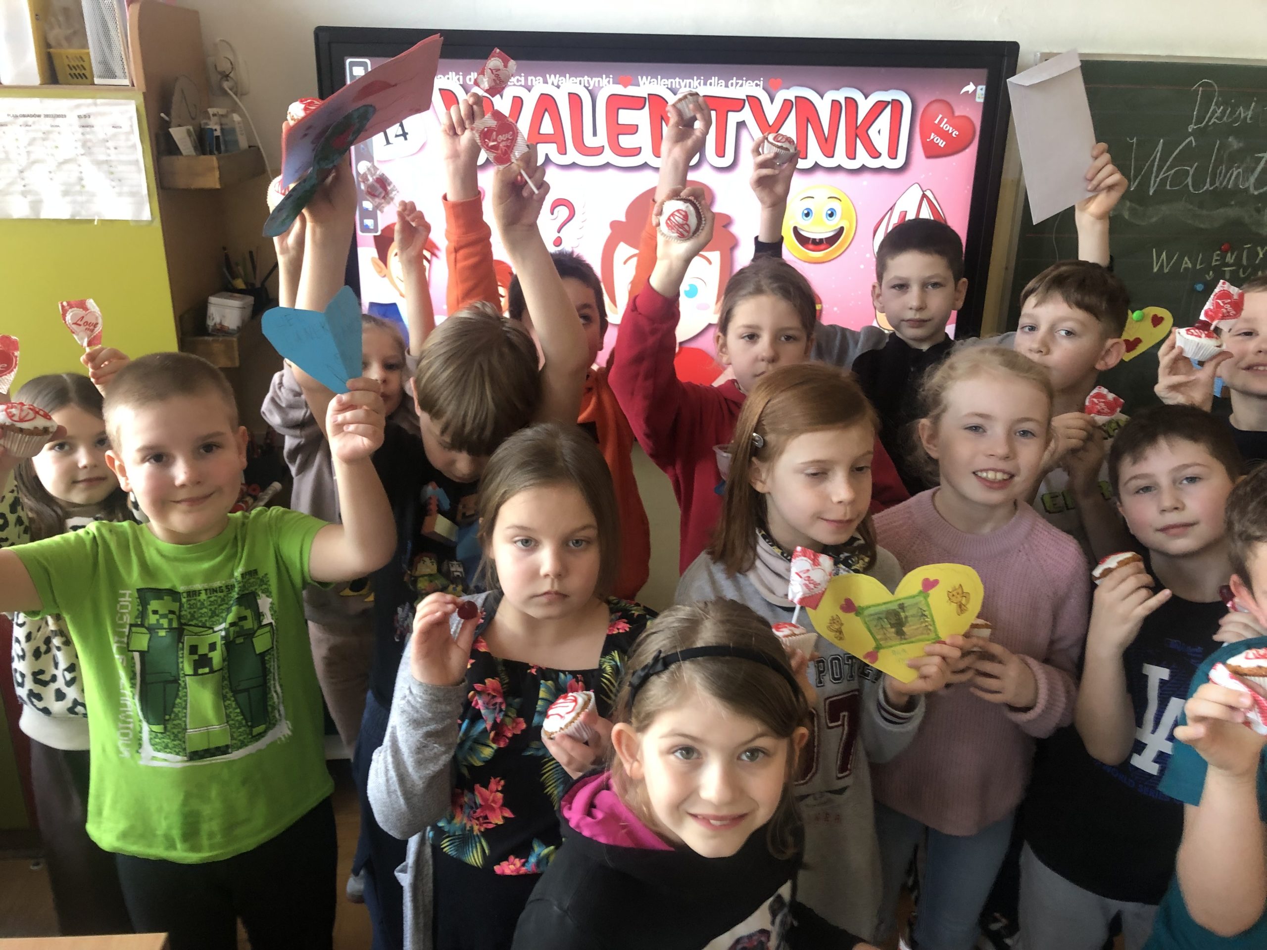 14 dzieci stoi na tle ekranu z napisem Walentynki. W rękach trzymają kartki walentynkowe, babeczki i lizaki.