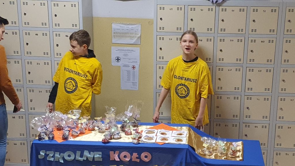 Dwoje uczniów Szkolnego Koła Wolontariatu Ustawionych w rzędzie, przed nimi stół na którym są wyłożone babeczki i ciastka