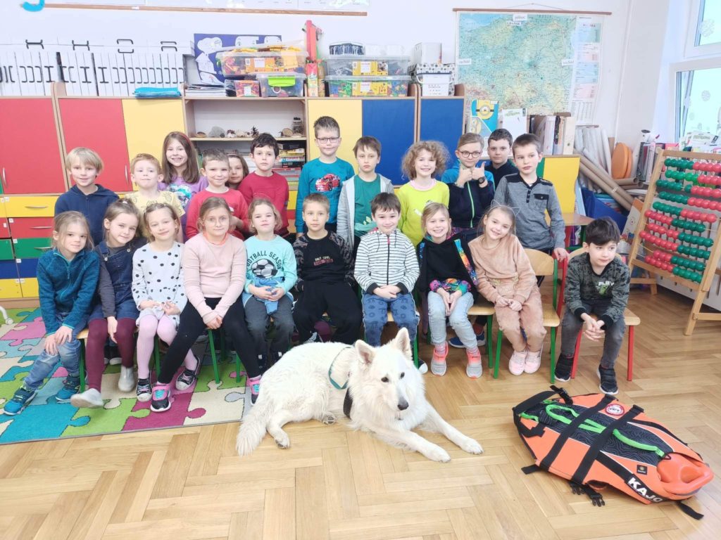 Grupka dzieci z psem ratownikiem w sali lekcyjnej na tle szfek szkonych