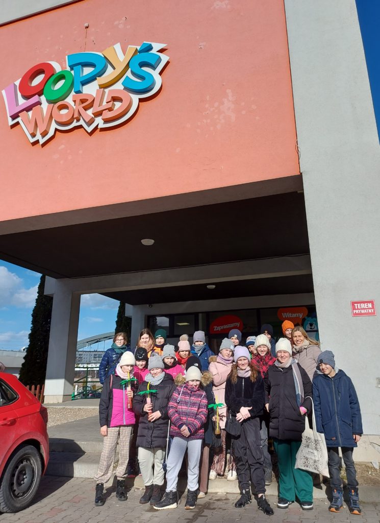 Grupa kilkunastu nastolatków ubranych w kurtki i czapki stoi przed budynkiem. Nad nimi jest napis Loopy’s World.