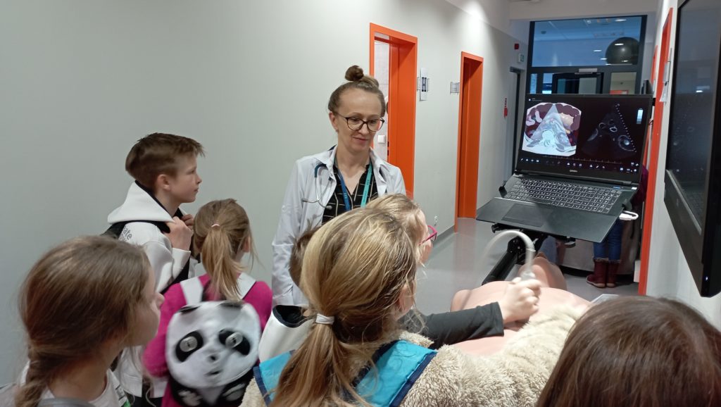 Na holu przy lekarce ubranej w biały fartuch stoi 7 uczniów. Jedna z uczennic trzyma głowicę aparatury USG na klatce piersiowej fantoma, a na monitorze laptopa wyświetla się obraz USG.