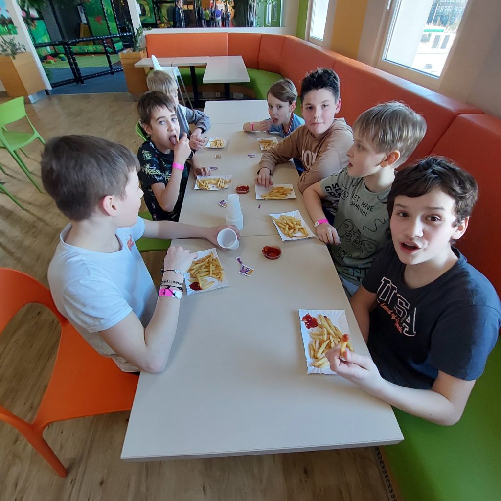 Siedmiu chłopców siedzi w restauracji przy stole, na którym są tacki z frytkami, pojemniczki z ketchupem i jednorazowe kubeczki.