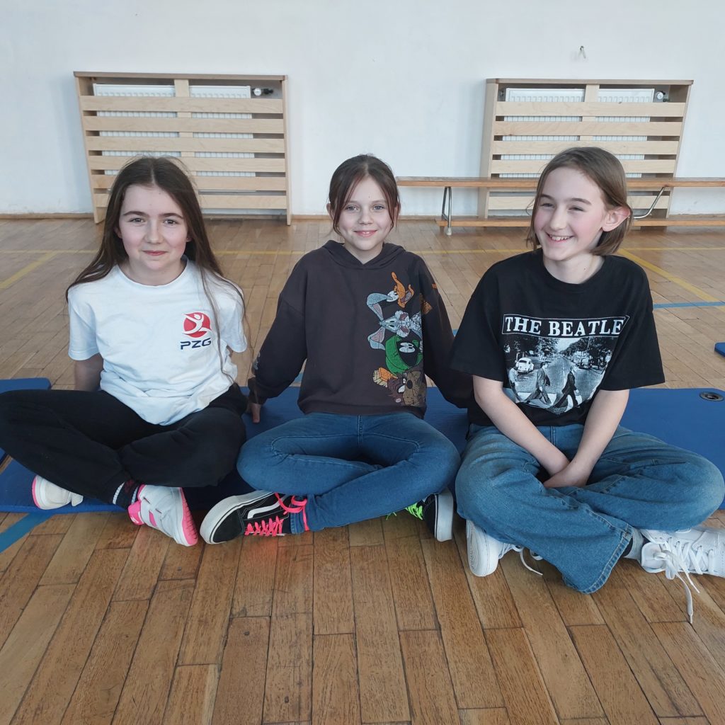 Trzy uśmiechnięte dziewczynki siedzą obok siebie na niebieskich matach. W tle jest sala gimnastyczna.