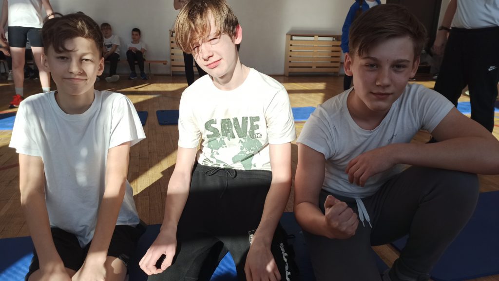 Trzech chłopców ubranych w białe koszulki siedzi obok siebie. W tle są uczniowie.