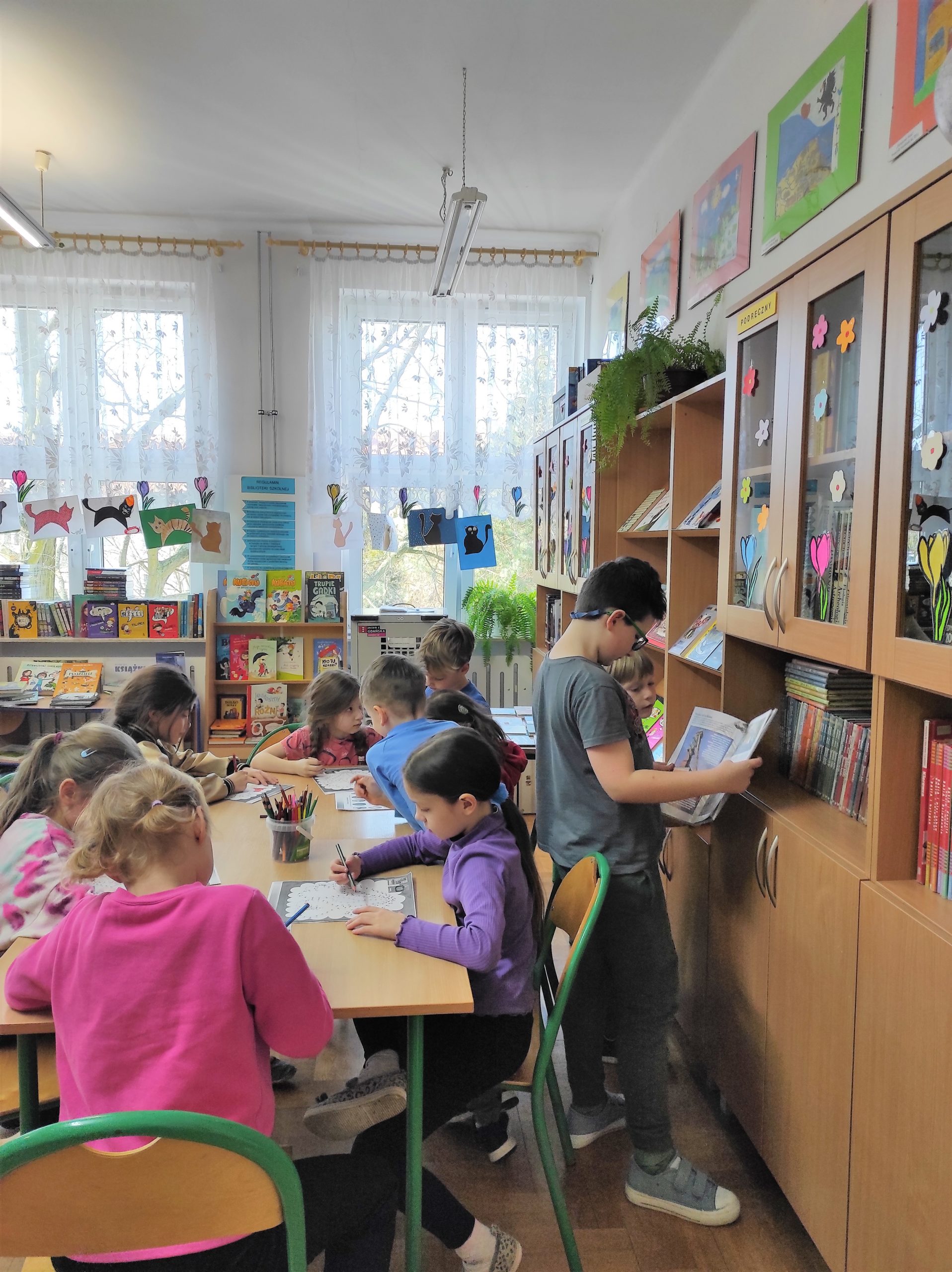 9 dzieci klas młodszych w czytelni biblioteki szkolnej, 3 uczniów czyta książkę, pozostałe dzieci rozwiązują rebusy
