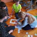 4 dzieci siedzi na podłodze w klasie. Wokół rozsypane puzzle z liniami. Dziewczynka po prawej i chłopiec po lewej trzymają ręce na puzzlach. Chłopiec po prawej trzyma w ręku świecącego się robota.
