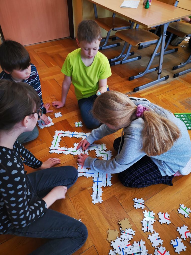 4 dzieci siedzi na podłodze w klasie. Wokół rozsypane puzzle z liniami. Dziewczynka po prawej i chłopiec po lewej trzymają ręce na puzzlach. Chłopiec po prawej trzyma w ręku świecącego się robota.